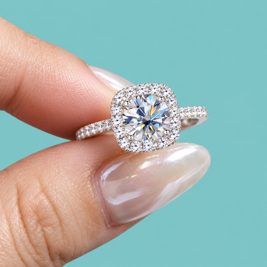 A close-up of a hand with manicured nails holding a silver halo ring featuring a cushion-cut diamond center stone and small accent diamonds on the band, set against a solid turquoise background.