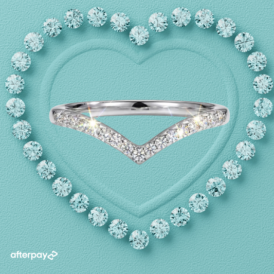 A silver V-shaped ring set with clear pavé stones, centered on a textured light teal background with a heart outline made of round clear stones. An Afterpay logo appears in the bottom-left corner.