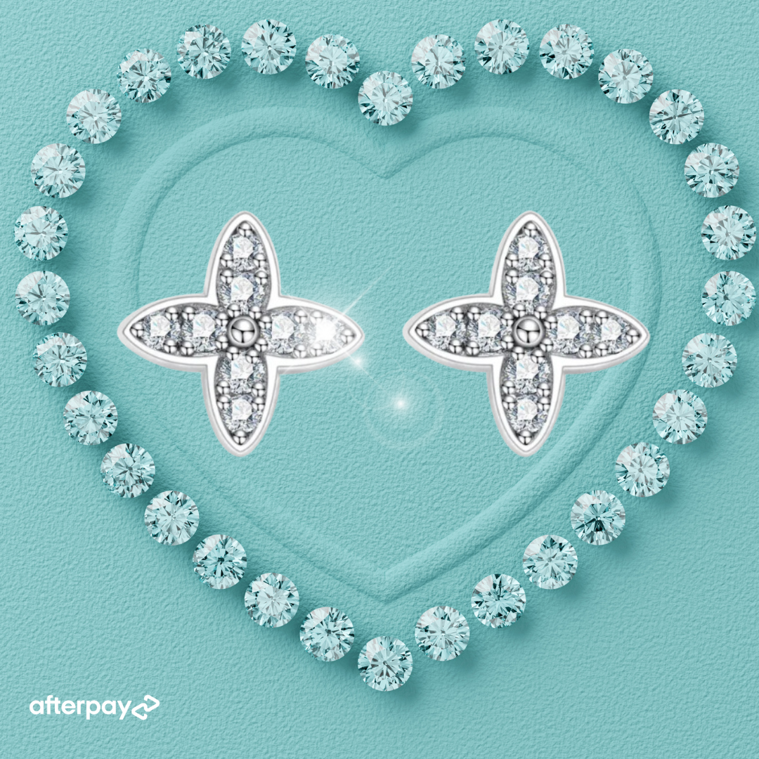 A pair of petal fleur de lis shape moissanite diamond earrings with a heart-shaped design, surrounded by smaller clear crystals, set on a teal background with a textured surface.