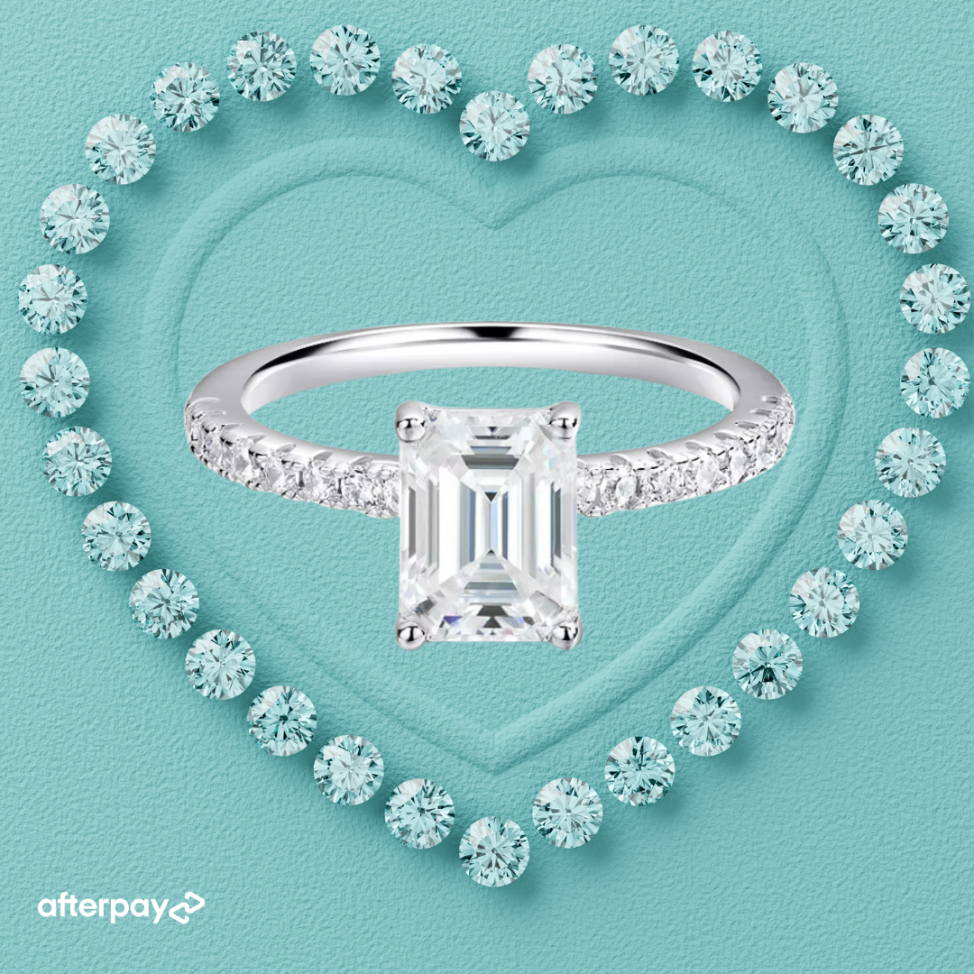 An emerald-cut moissanite solitaire ring with a pavé band, displayed on a heart-shaped background.
