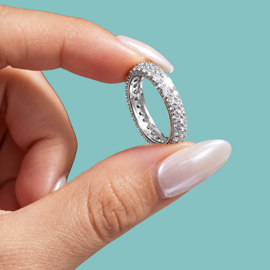 A close-up of a hand with long, manicured nails holding a silver eternity ring paved with small clear stones against a solid teal background.