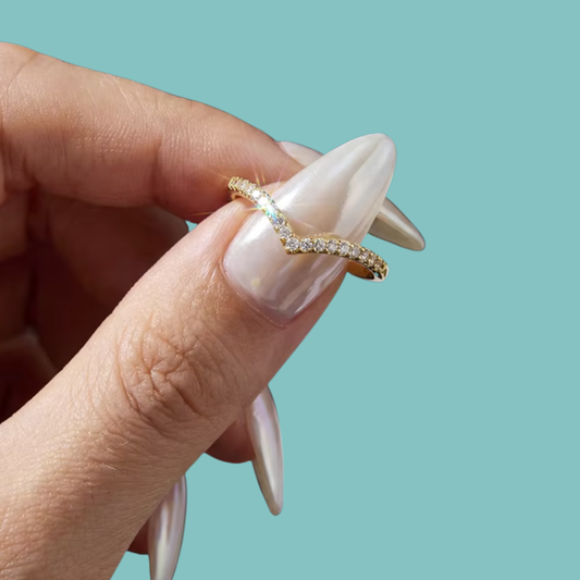 A close-up of a hand with long, glossy nude manicured nails holding a thin wavy gold ring adorned with small clear stones against a solid light teal background.