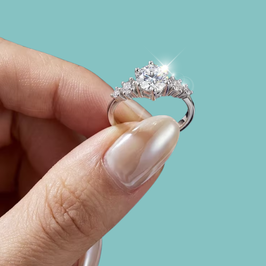 A close-up of a hand holding a silver-toned ring with a sparkling clear center stone and smaller side stones, set against a solid light aqua background.