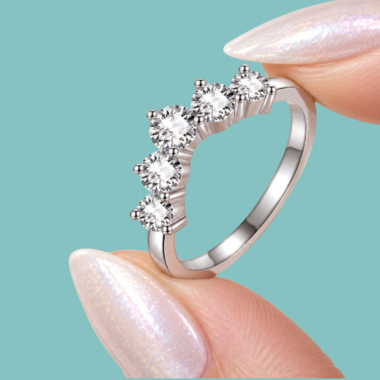 A silver-toned ring with five clear round-cut stones arranged in a curved row, held between two manicured fingernails against a solid light aqua background.