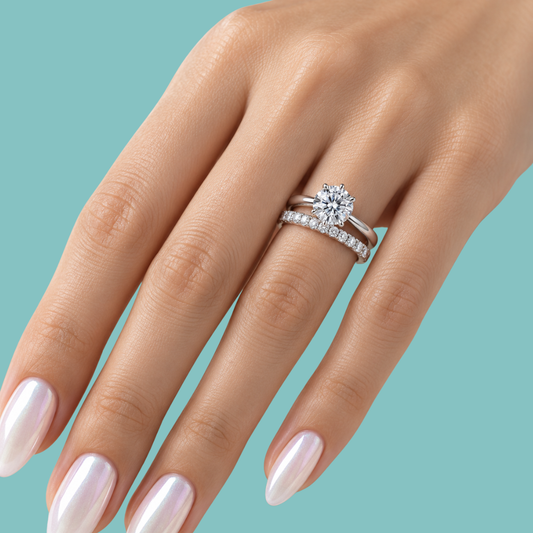 A close-up of a hand wearing a silver engagement ring with a round-cut center stone and a matching diamond band, set against a solid teal background.