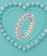 A silver eternity ring set with clear round-cut gemstones, presented on a textured turquoise background with a heart outline formed by matching stones.