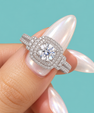 A close-up of a hand with long, glossy pale pink nails wearing a silver-toned halo ring with a large round clear center stone and smaller surrounding stones, set against a solid light aqua background.