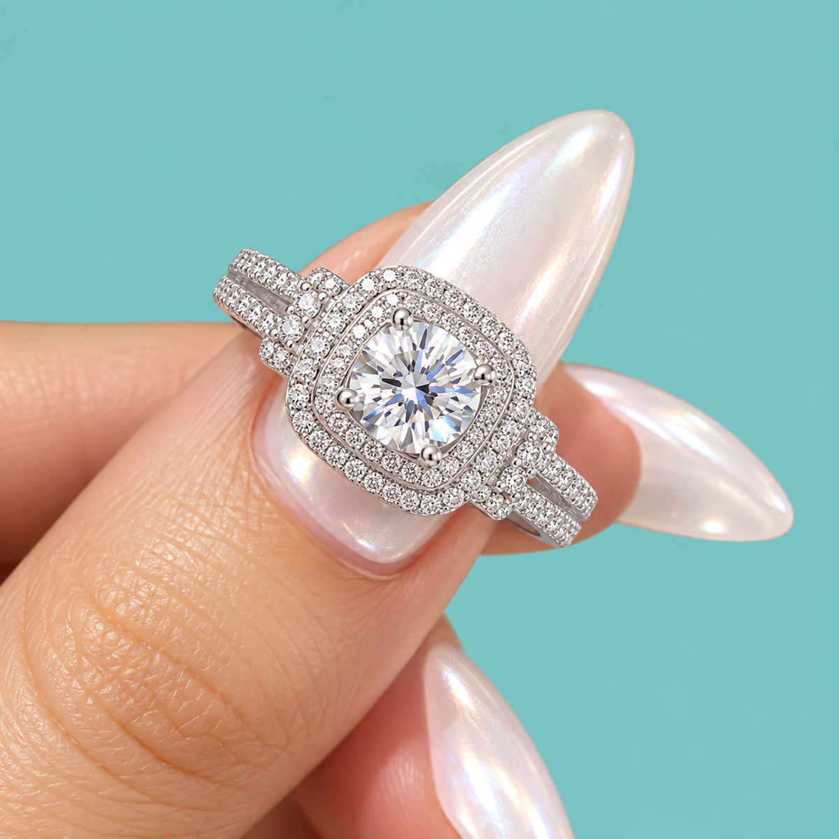 A close-up of a hand with long, glossy pale pink nails wearing a silver-toned halo ring with a large round clear center stone and smaller surrounding stones, set against a solid light aqua background.