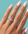 A close-up of a hand with long, glossy white manicured nails wearing a silver engagement ring with a round brilliant-cut center stone and a matching pave band, set against a solid light blue background.