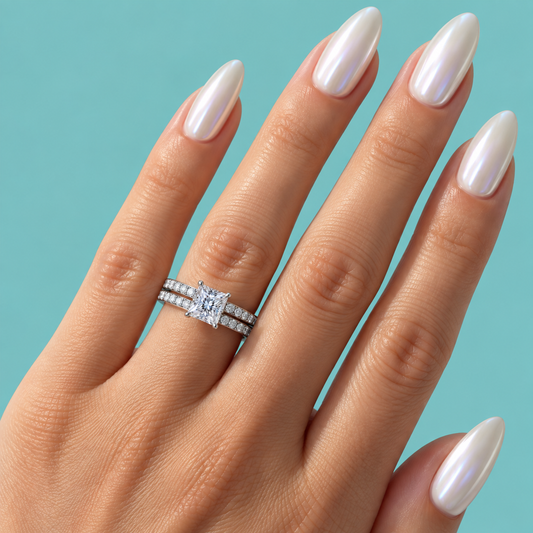 A close-up of a hand with long, glossy white manicured nails wearing a silver engagement ring with a round brilliant-cut center stone and a matching pave band, set against a solid light blue background.