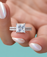 A close-up of a hand with iridescent white nail polish holding a silver engagement ring featuring a princess-cut clear center stone and pavé-set band, against a solid teal background.