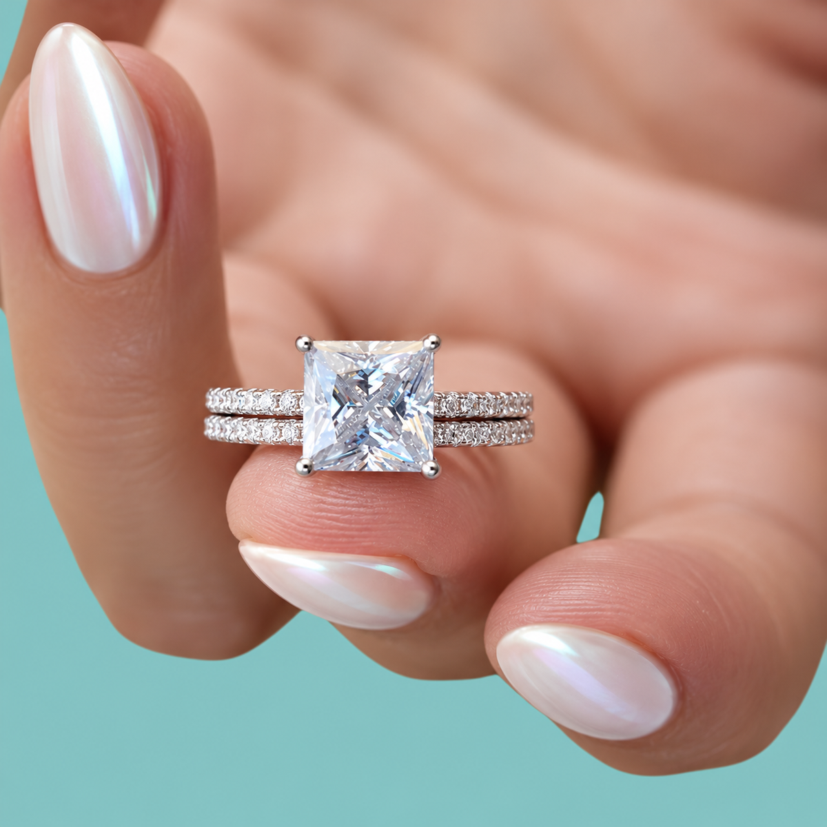 A close-up of a hand with iridescent white nail polish holding a silver engagement ring featuring a princess-cut clear center stone and pavé-set band, against a solid teal background.