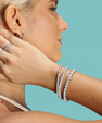 A close-up lifestyle shot of a woman wearing a silver ring with a square-cut gemstone on her finger, a silver hoop earring, and three silver tennis bracelets on her wrist, set against a solid teal background.