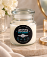 A clear glass jar candle with white wax sits on a marble surface in a styled indoor setting. The jar has a black circular label with white and blue text reading 'FIRE AND MOONLIGHT looks just like a diamond but better!' and small ring graphics.