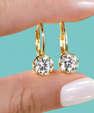 A close-up of a person's hand holding a pair of gold hoop earrings with round clear gemstones against a solid turquoise background.