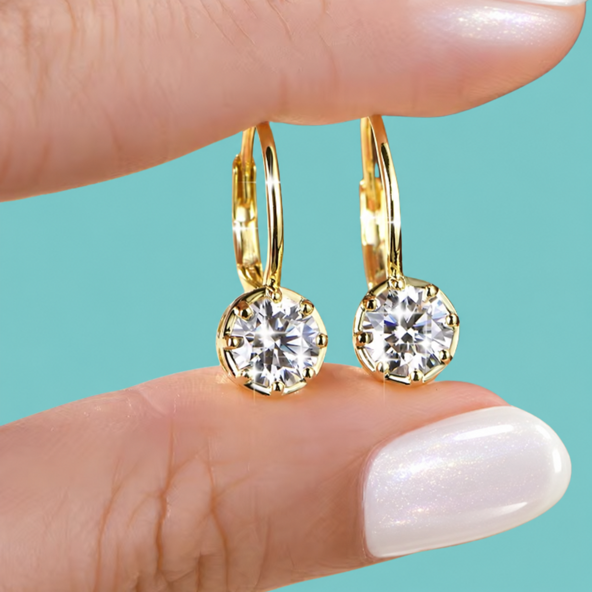 A close-up of a person's hand holding a pair of gold hoop earrings with round clear gemstones against a solid turquoise background.