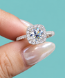A close-up of a hand with manicured nails holding a silver halo ring featuring a cushion-cut diamond center stone and small accent diamonds on the band, set against a solid turquoise background.