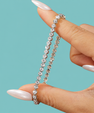 A close-up of a hand with long, glossy nude manicured nails holding a silver tennis bracelet with clear round-cut stones against a solid teal background.