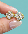 A pair of gold flower-shaped stud earrings with clear round-cut stones, held between two fingers against a solid turquoise background.