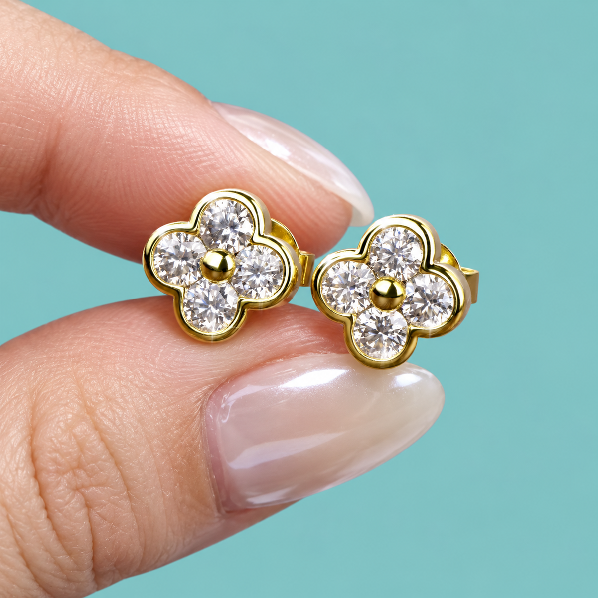A pair of gold flower-shaped stud earrings with clear round-cut stones, held between two fingers against a solid turquoise background.