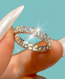 A close-up of a silver diamond eternity ring held between fingers with long, glossy white manicured nails, set against a solid turquoise background.