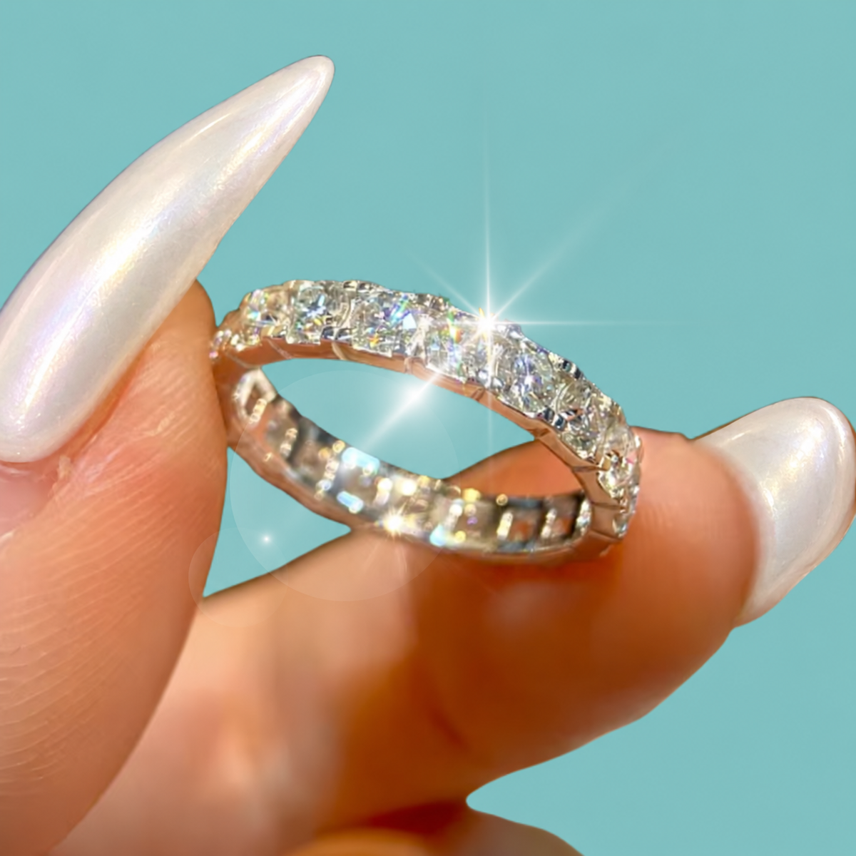 A close-up of a silver diamond eternity ring held between fingers with long, glossy white manicured nails, set against a solid turquoise background.