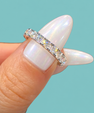 A close-up of a diamond eternity ring on a finger with long, iridescent white manicured nails, set against a solid light blue background.