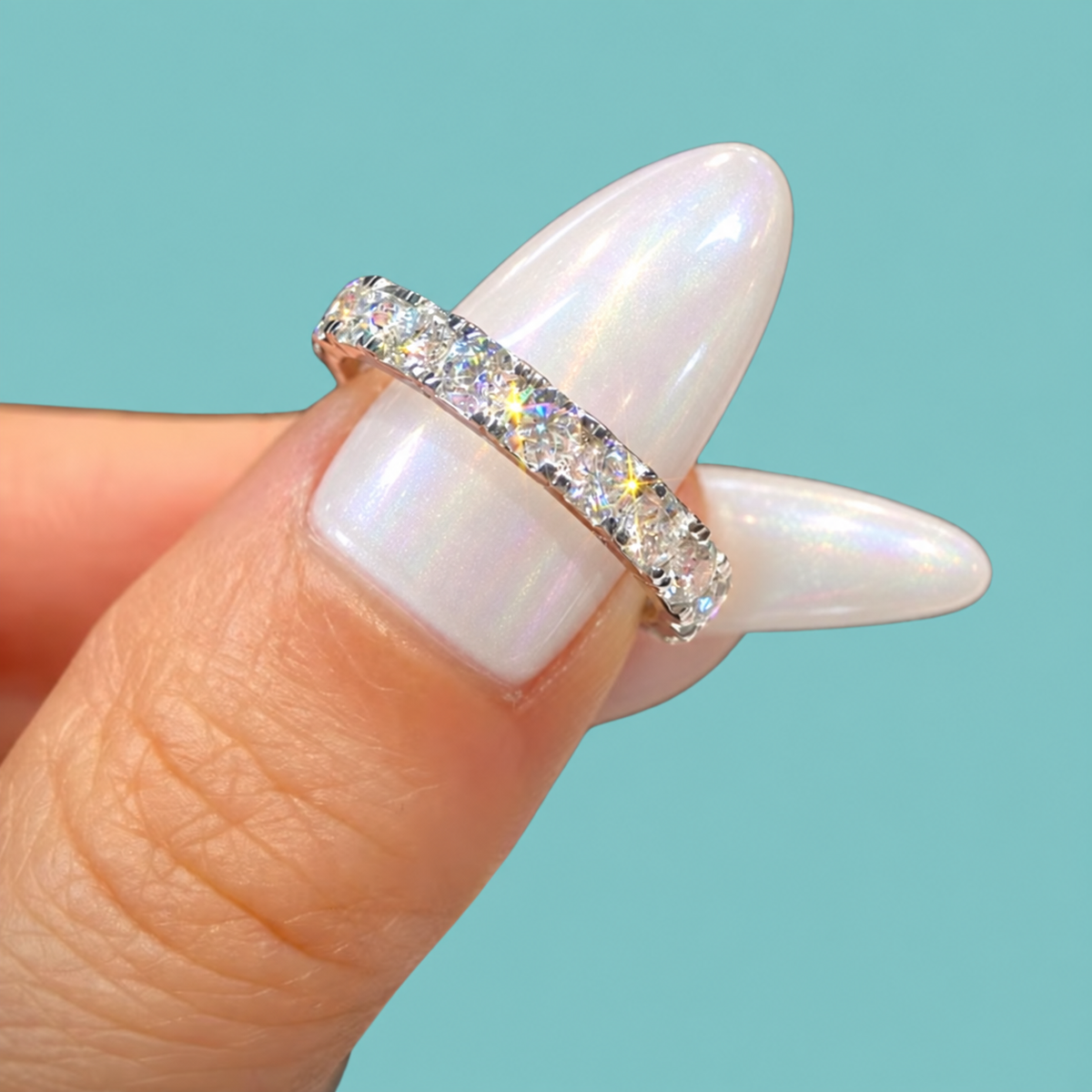A close-up of a diamond eternity ring on a finger with long, iridescent white manicured nails, set against a solid light blue background.