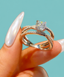 A close-up of a hand with long, glossy white manicured nails holding a double-band engagement ring. The ring features a brilliant round-cut clear center stone on a silver-toned band with pavé-set accent stones, set against a solid light teal background.