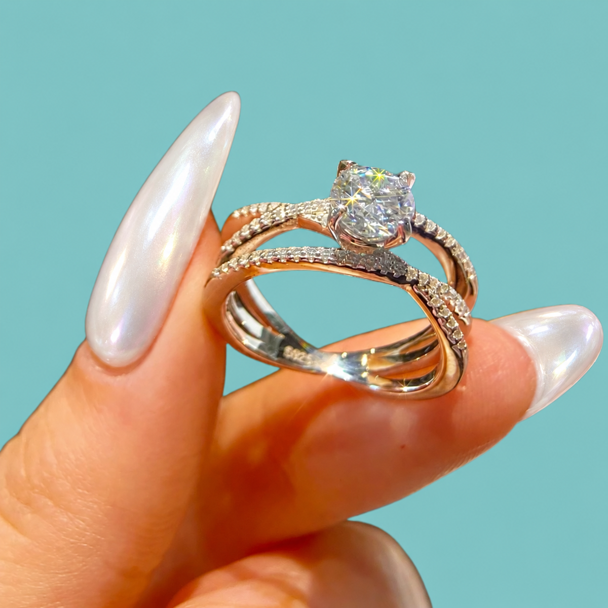 A close-up of a hand with long, glossy white manicured nails holding a double-band engagement ring. The ring features a brilliant round-cut clear center stone on a silver-toned band with pavé-set accent stones, set against a solid light teal background.