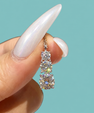 A close-up of a hand with long, glossy, iridescent white fingernails holding a small gold triple-stone diamond pendant. The background is a solid, clean turquoise color.