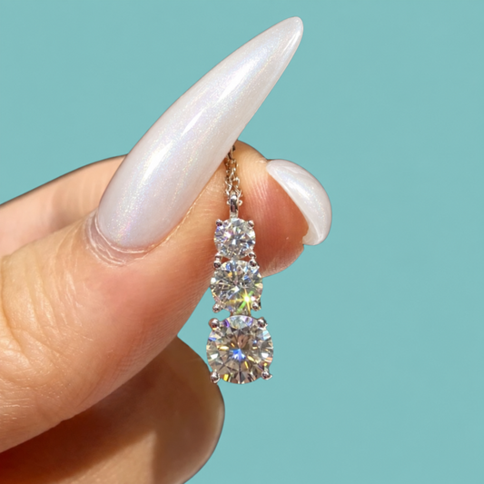 A close-up of a hand with long, glossy, iridescent white fingernails holding a small gold triple-stone diamond pendant. The background is a solid, clean turquoise color.