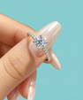 A close-up of a hand with long, manicured nails holding a silver-toned engagement ring with a round-cut center stone and a pavé band, set against a solid light turquoise background.