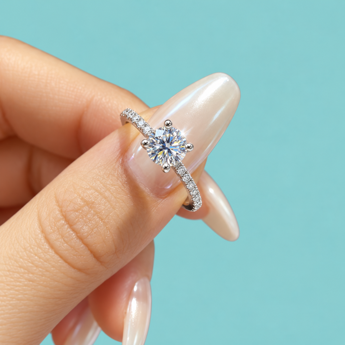 A close-up of a hand with long, manicured nails holding a silver-toned engagement ring with a round-cut center stone and a pavé band, set against a solid light turquoise background.