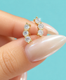 A close-up of a hand with long, manicured nails holding a pair of gold huggie hoop earrings set with clear round-cut gemstones, against a solid light blue background.