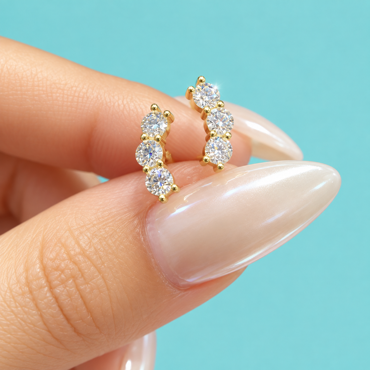 A close-up of a hand with long, manicured nails holding a pair of gold huggie hoop earrings set with clear round-cut gemstones, against a solid light blue background.