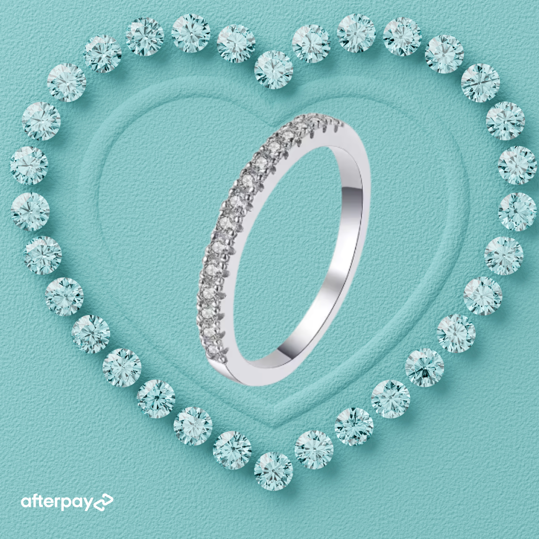 A silver-toned eternity ring set with round brilliant-cut diamonds, presented on a textured light blue background with a heart-shaped border of matching diamonds. The Afterpay logo is visible in the bottom-left corner.