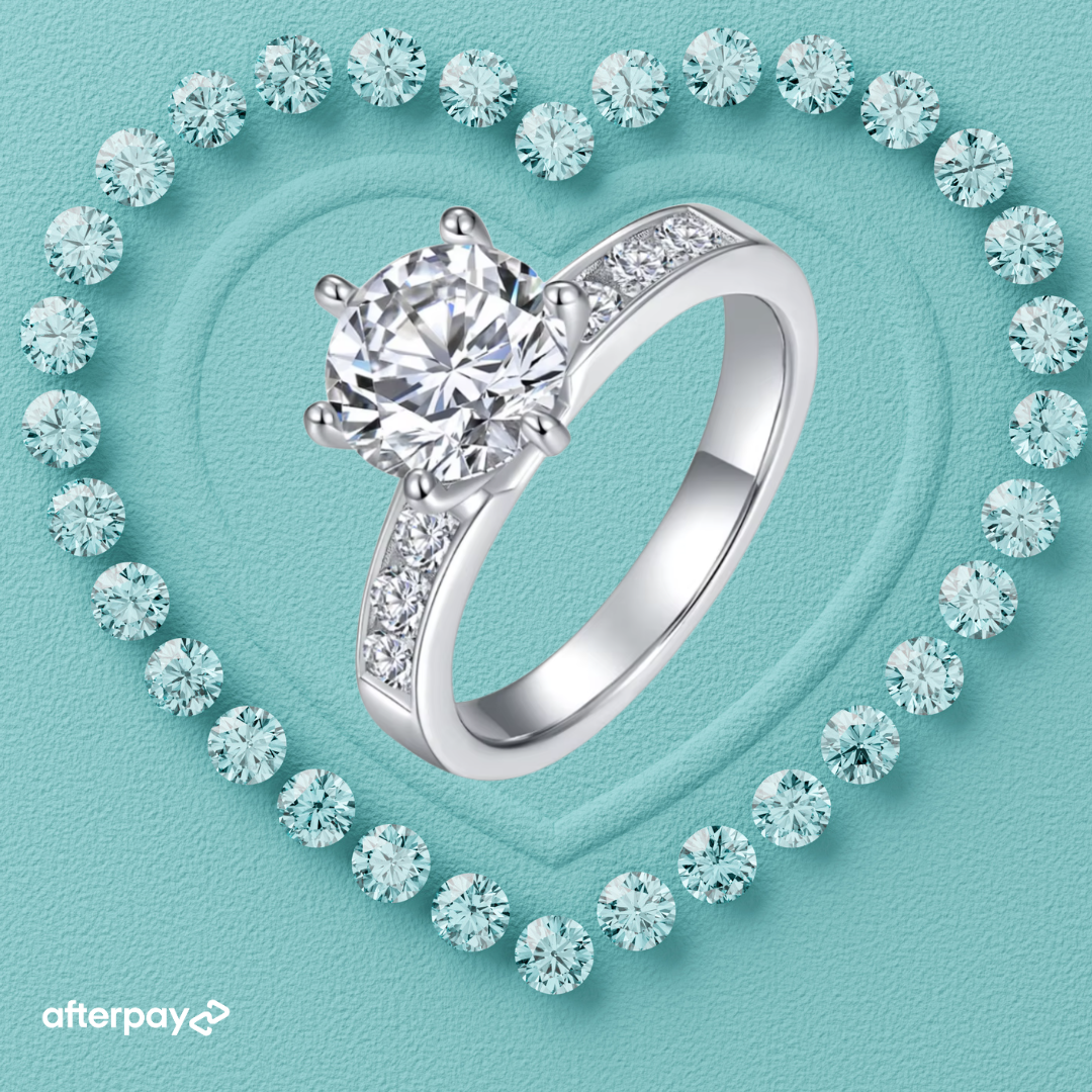 A silver moisasnite 2 carat engagement ring with a large round-cut diamond center stone and smaller diamonds along the band, displayed on a turquoise heart-shaped background surrounded by small diamond-like stones. The Afterpay logo appears in the lower-left corner.