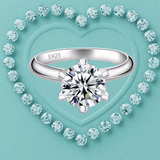 A silver solitaire engagement ring with a round brilliant-cut diamond, featuring an 'S925' hallmark on the inner band. The ring is centered against a textured light blue background, framed by a heart-shaped border of small sparkling diamonds.