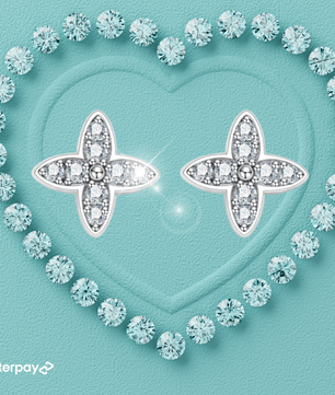 A pair of petal fleur de lis shape moissanite diamond earrings with a heart-shaped design, surrounded by smaller clear crystals, set on a teal background with a textured surface.