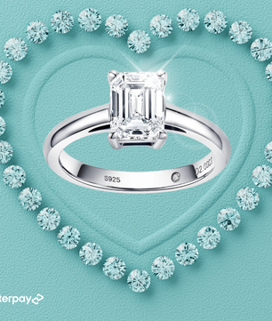 A 2 carat emerald cut solitaire moissanite ring with a classic polished band, surrounded by smaller moissanite stones, set on a heart-shaped inset on a turquoise background.