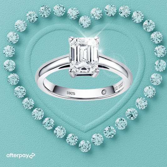 A 2 carat emerald cut solitaire moissanite ring with a classic polished band, surrounded by smaller moissanite stones, set on a heart-shaped inset on a turquoise background.