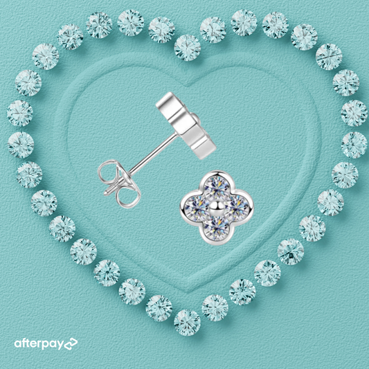A pair of silver stud earrings arranged on a light aqua textured background, with one earring shown front-facing as a four-petal flower and the other shown side-facing to reveal the post and butterfly back. The earrings are surrounded by a heart-shaped border of clear gemstones, and an Afterpay logo appears in the bottom-left corner.