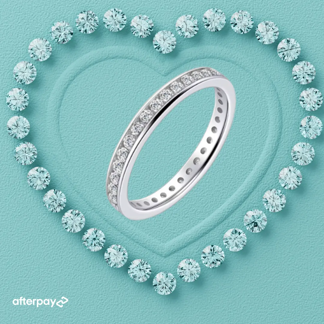 A silver-tone eternity ring set with clear round-cut gemstones, arranged on a textured aqua background with a heart outline made of matching stones. An Afterpay logo appears in the bottom-left corner.