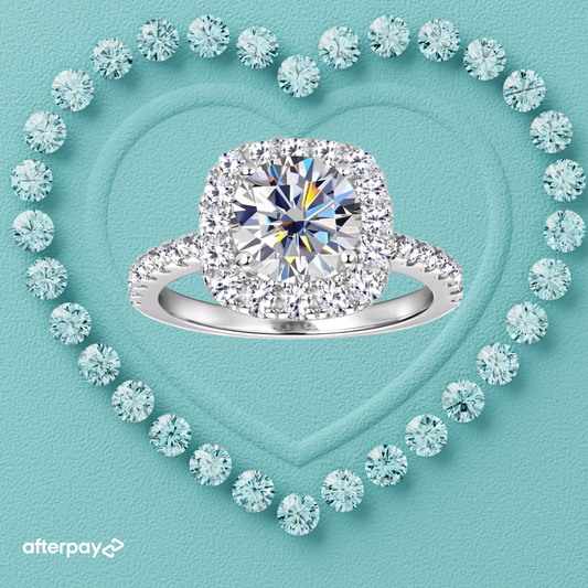 A double-halo diamond engagement ring with a silver-colored band, centered on a textured light blue background with a heart outline made of smaller diamonds. An Afterpay logo is visible in the bottom-left corner.