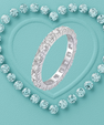 A silver eternity ring set with round brilliant-cut diamonds, presented on a textured light blue background with a heart-shaped border of matching diamonds.