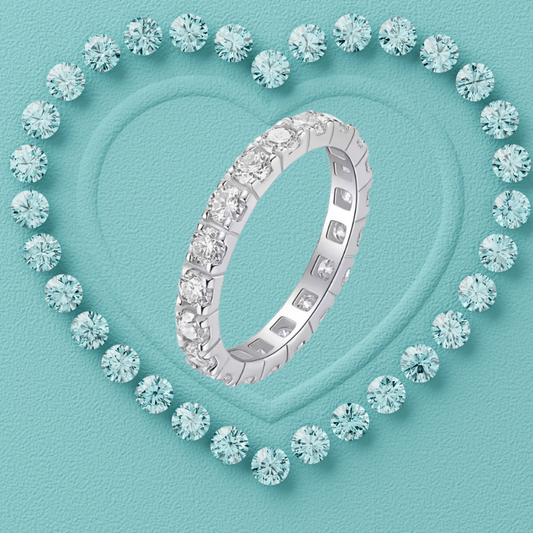 A silver eternity ring set with round brilliant-cut diamonds, presented on a textured light blue background with a heart-shaped border of matching diamonds.