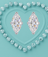 A pair of marquise-cut diamond stud earrings in a silver setting, arranged side-by-side in the center of a heart-shaped arrangement of round brilliant-cut diamonds on a textured light blue background.