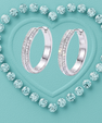 A pair of silver hoop earrings with pavé-set clear stones, arranged on a textured light aqua background with a heart outline made of matching round stones.