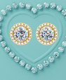 A pair of gold stud earrings with large round clear center stones surrounded by small clear stones, arranged inside a heart outline made of light blue gemstones on a turquoise background.